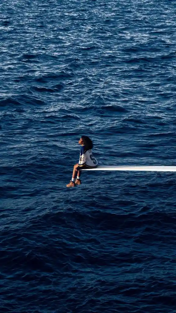 A person sits alone on a narrow white plank suspended over a vast, deep blue ocean, looking away from the camera. The person…