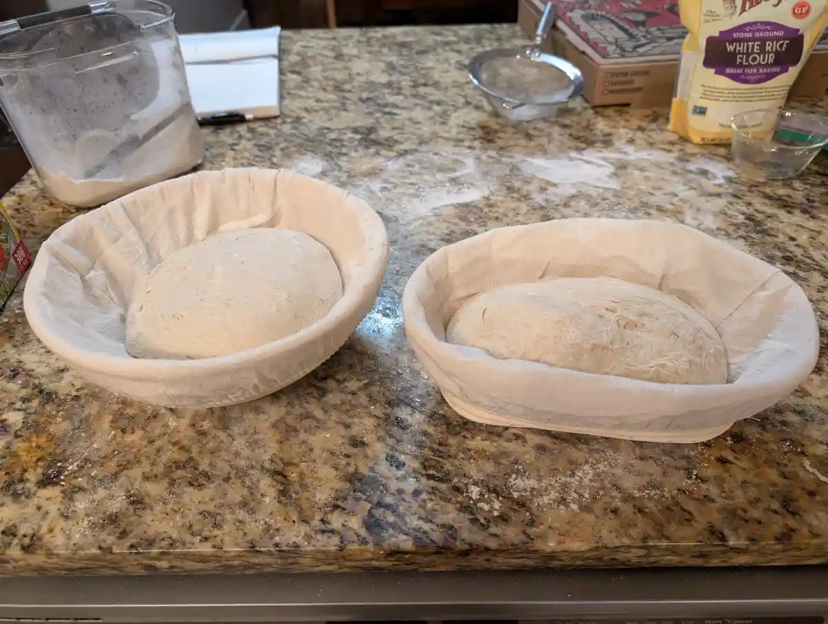 Two balls of dough sit in cloth-lined baskets on a flour-dusted granite countertop, one round and one lopsided. The overlaid…