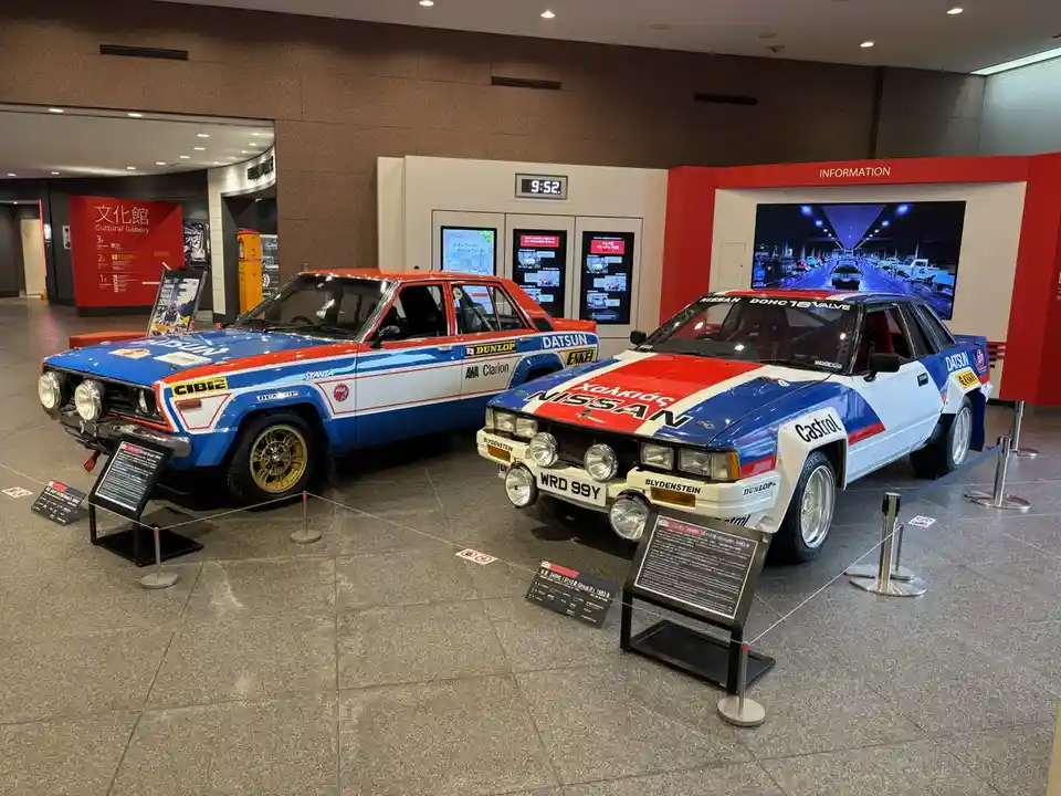 Two vintage Japanese rally cars, a blue, red, and white Datsun Stanza and a Nissan S110, are displayed side-by-side in a…