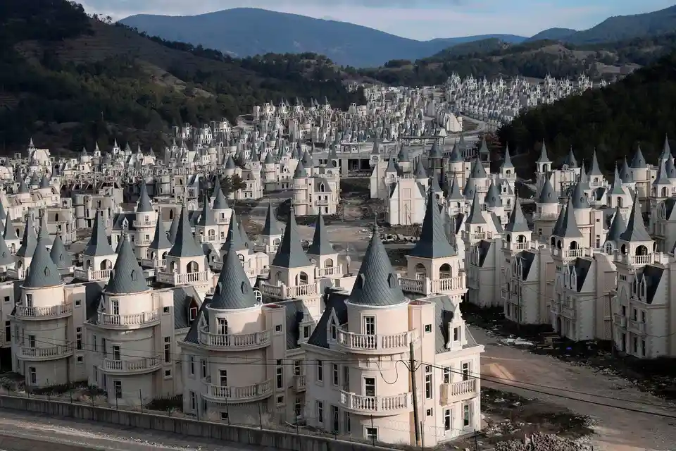 An aerial view shows a vast, unfinished residential development with hundreds of identical, castle-like houses featuring…
