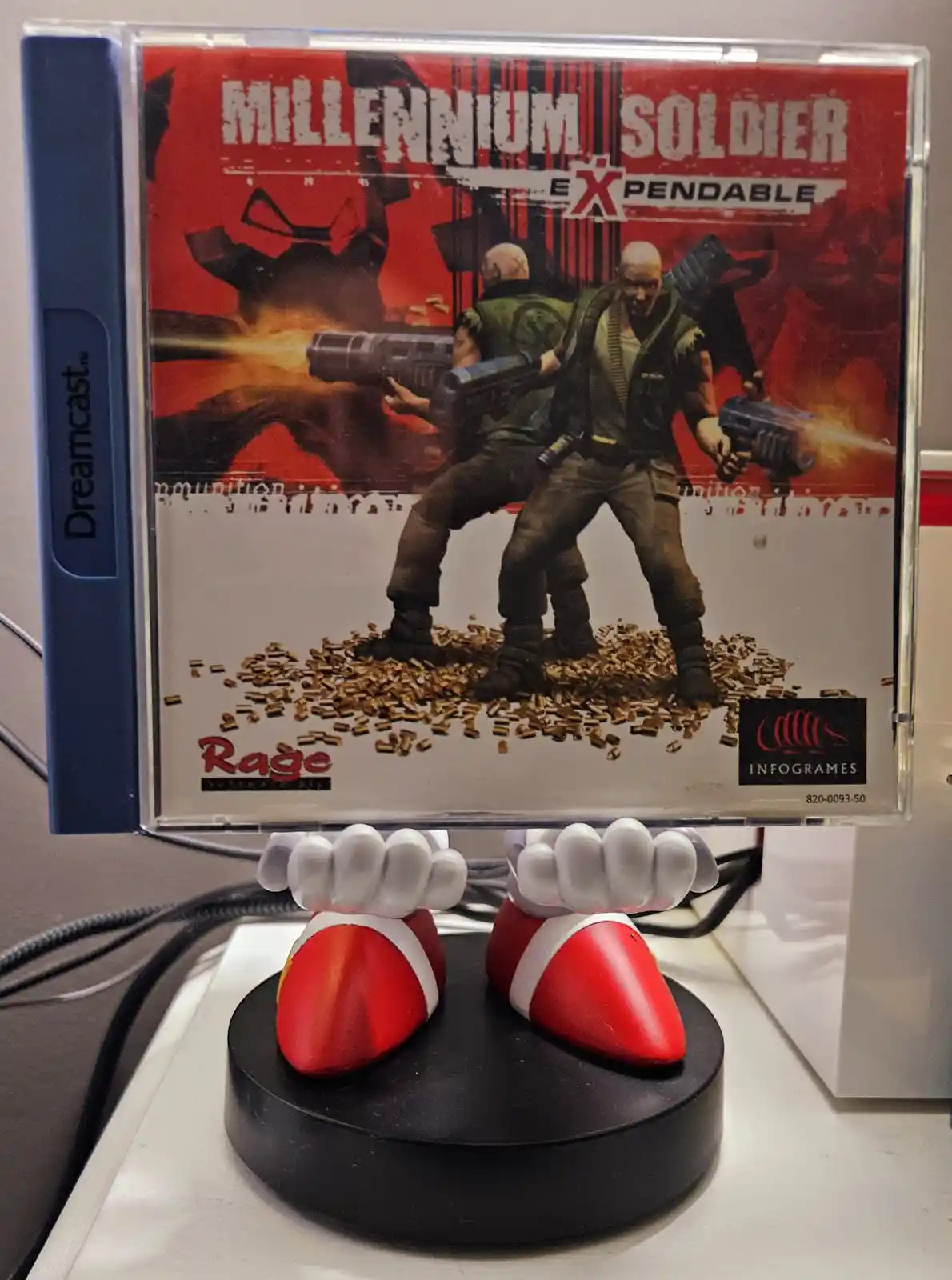 A Dreamcast game case for 'Millennium Soldier Expendable' is propped up by a figurine of Mario's feet in red and white…