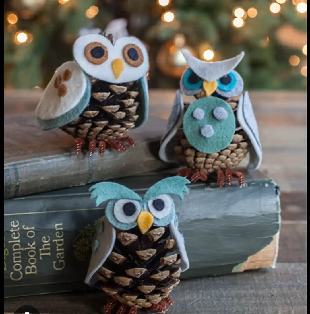 Three handmade owl decorations, crafted from pinecones with felt faces and wings, perch atop stacked books. The background…