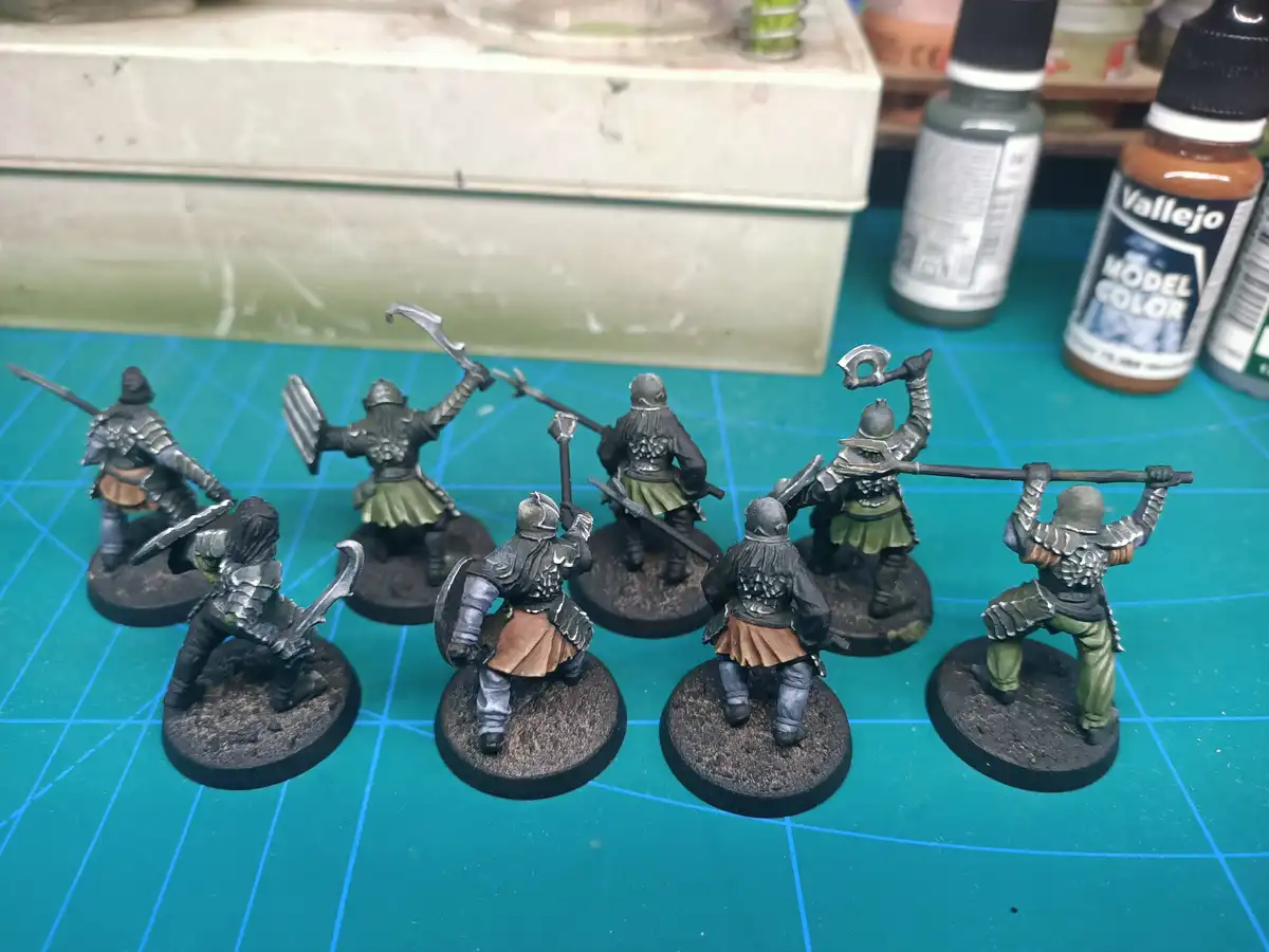 A photo shows seven painted fantasy miniatures, likely dwarves or warriors, standing on circular bases on a green grid mat…