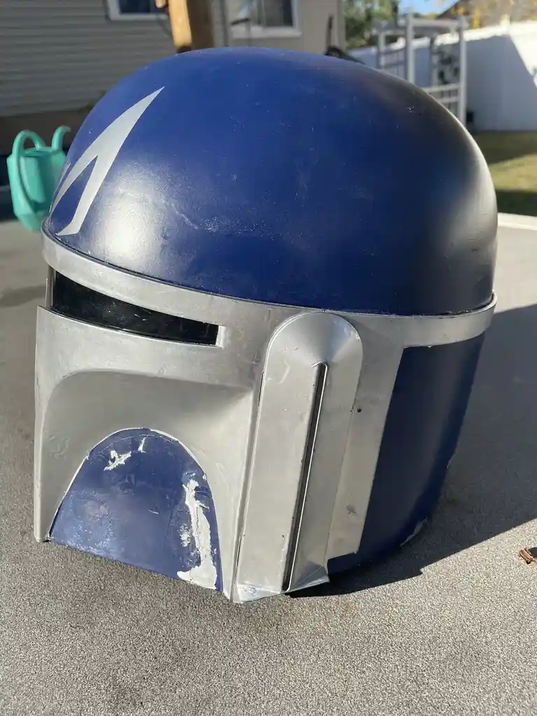 A blue and silver Mandalorian-style helmet with a white lightning bolt on top sits on a paved surface outdoors, showing…