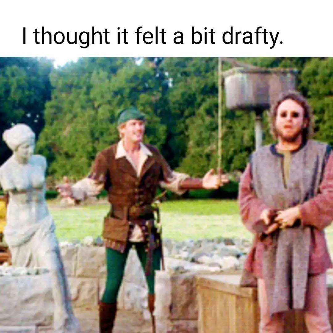 Two men in medieval-style clothing stand outdoors near a stone statue and wooden structures. The man on the left, wearing a…
