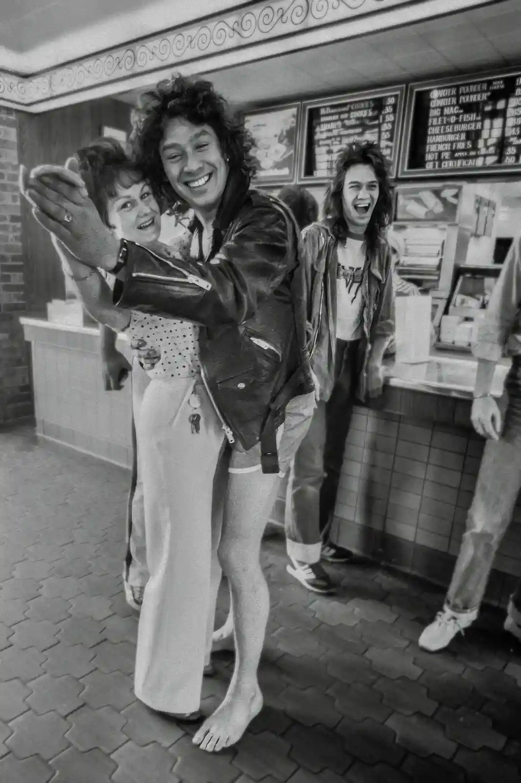 A black-and-white photo shows a young man in a leather jacket and shorts, barefoot on a tiled floor, hugging a woman while…