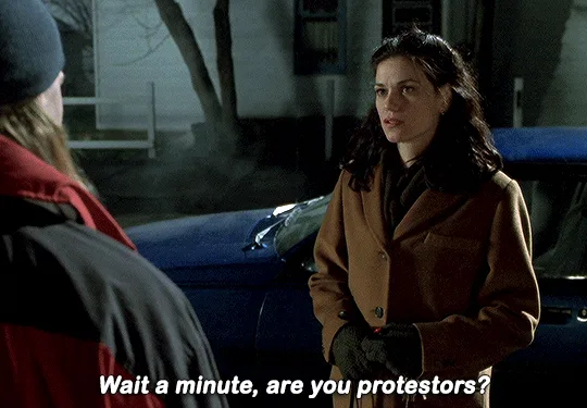 The image shows a woman in a brown coat and black gloves standing outdoors at night, facing a person in a red and black…