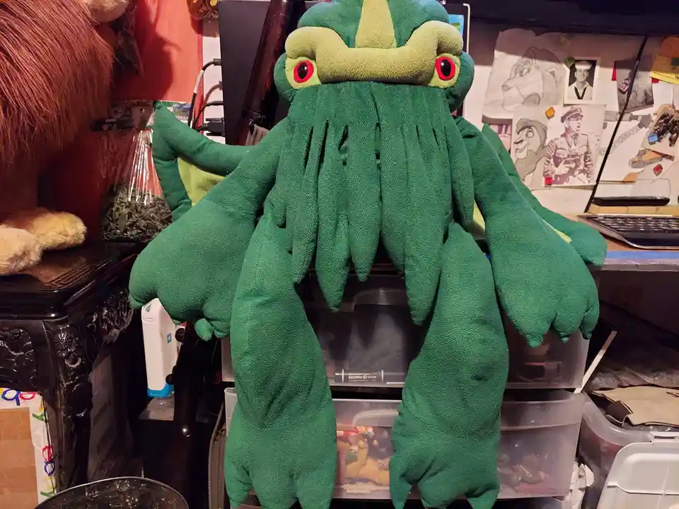 A large green plushie resembling a Cthulhu-like monster with multiple tentacles and red eyes sits on a cluttered desk…