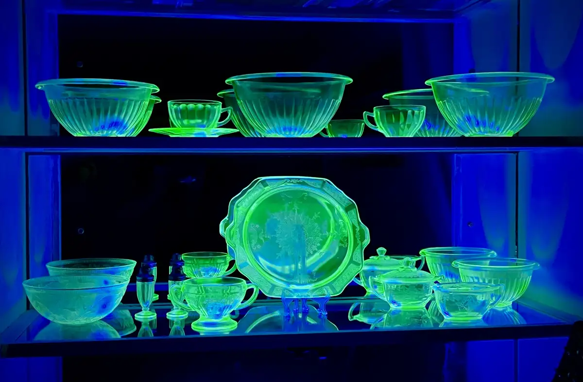 A display case under blacklight shows various pieces of uranium glassware, including bowls, cups, and a large decorative…
