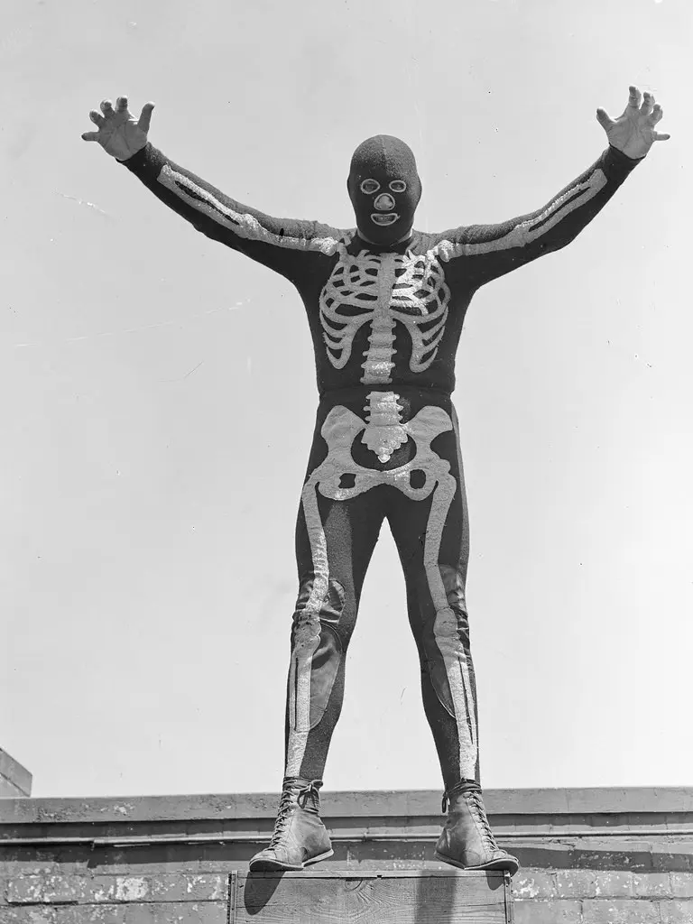 A black-and-white photo shows a person in a full-body skeleton costume standing atop a wooden platform with arms raised. The…