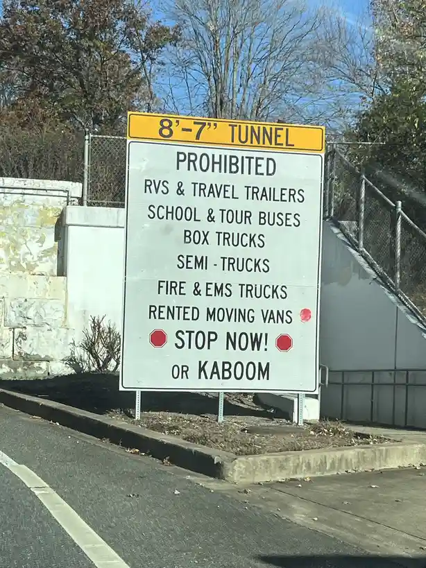 A yellow and white road sign warns drivers that an 8'7" tunnel prohibits RVs, buses, box trucks, semi-trucks, fire/EMS…