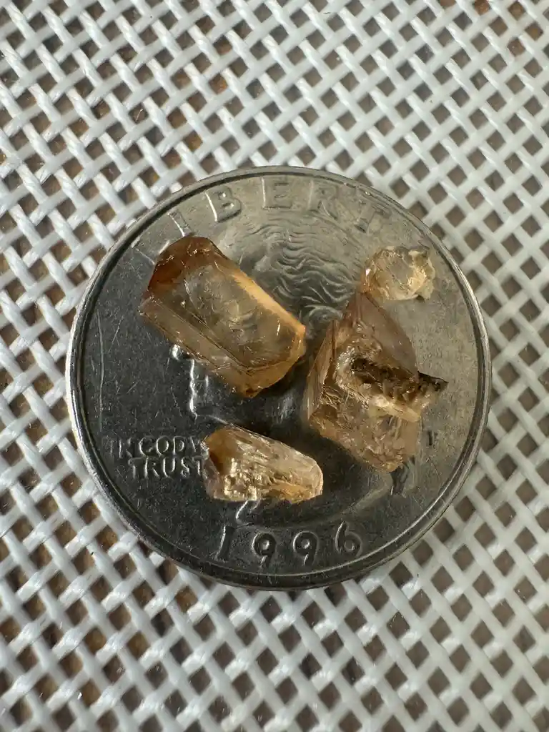 A close-up photo shows four small, translucent brownish crystals resting on a 1996 U.S. quarter, which is placed on a white…