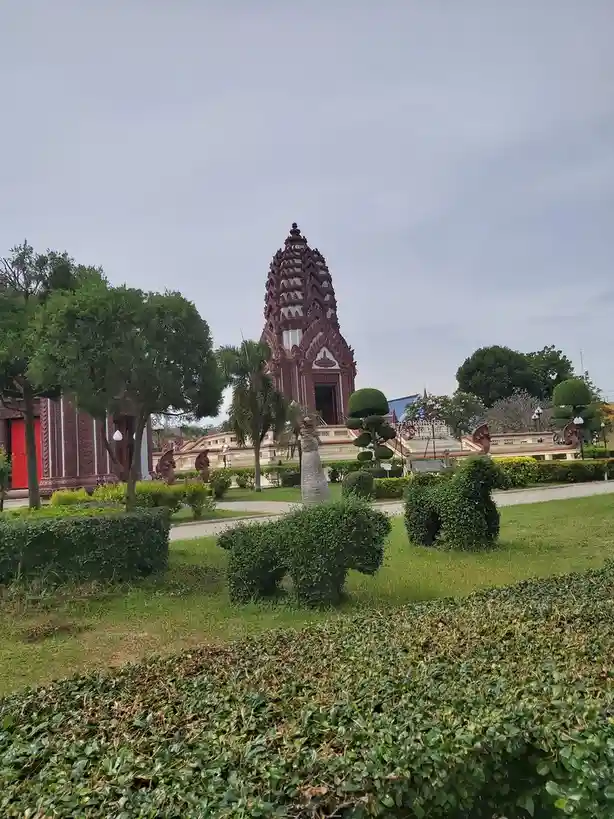 A photo of a red and white Thai temple with ornate architecture, surrounded by manicured green hedges and trees under an…