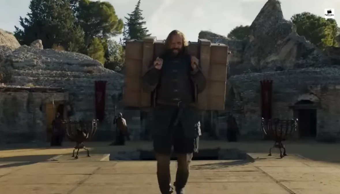 A bearded man in medieval-style clothing walks forward while carrying a large, cumbersome stack of cardboard boxes on his…