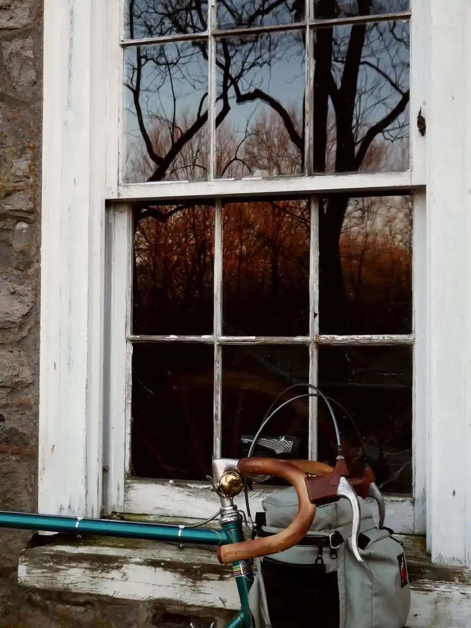 A teal bicycle with wooden handlebars and a grey bag rests against a weathered white window frame. The window reflects bare…