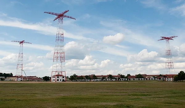Three tall, red lattice towers resembling giant cranes stand in a grassy field under a partly cloudy sky, with a row of low…