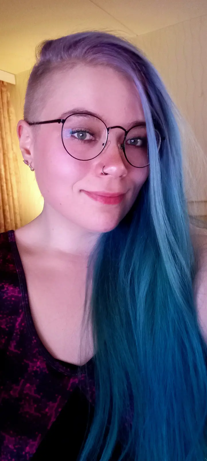 A person with a shaved head on one side and long, vibrant blue-green hair on the other, wearing round glasses and a nose…