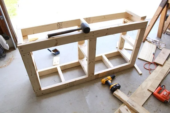 A partially assembled wooden frame sits on a concrete floor in a garage, with a hammer resting across the top. Tools like a…
