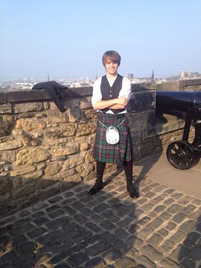A young man with brown hair stands confidently on a stone wall with arms crossed, wearing a traditional Scottish kilt, white…