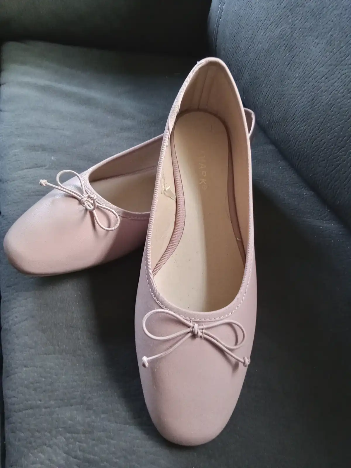 A pair of light pink ballet flats with bows on the toes sits on a dark gray textured couch. The shoes are positioned with…