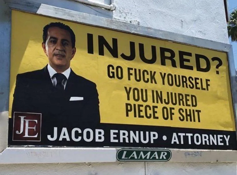A yellow billboard features a man in a suit with the text 'INJURED? GO FUCK YOURSELF YOU INJURED PIECE OF SHIT' above his…