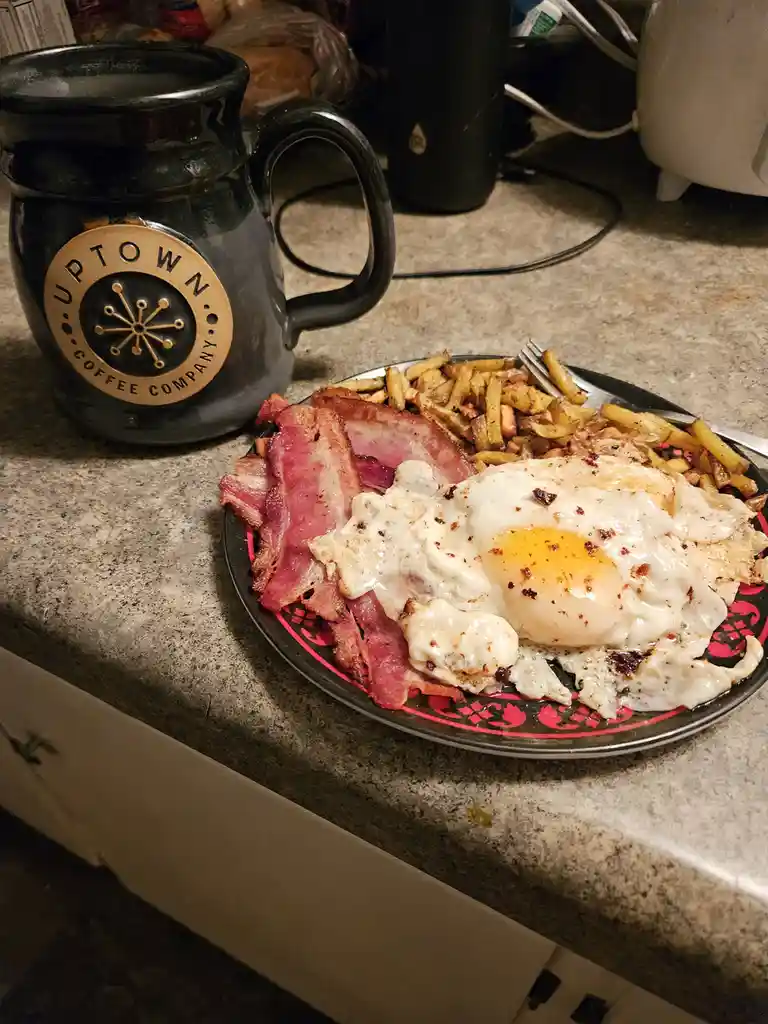 A plate of breakfast food including bacon, a fried egg, and french fries sits on a kitchen counter next to a black mug with…