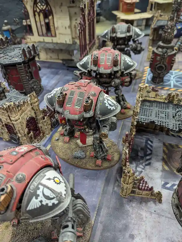 A photo of several large, red and grey painted sci-fi war machines, resembling heavily armored walkers or titans, positioned…