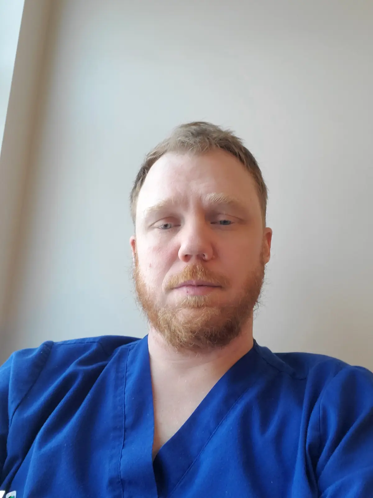 A man with a reddish beard and short hair, wearing blue medical scrubs, stares directly at the camera with a weary…
