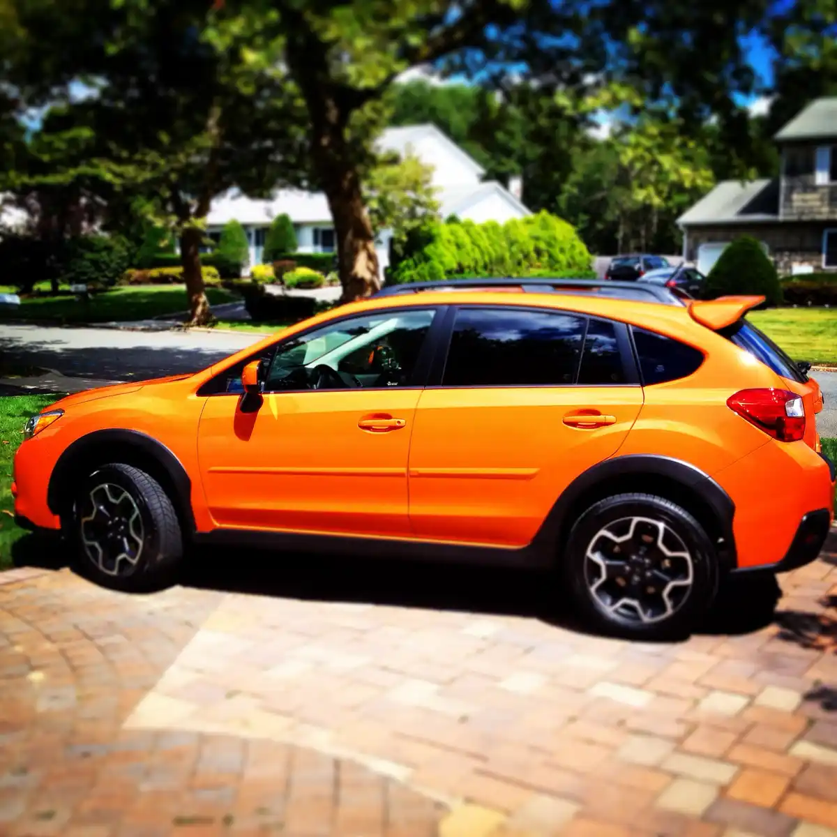 A bright orange Subaru Crosstrek SUV is parked on a brick driveway in a suburban neighborhood with houses and trees in the…