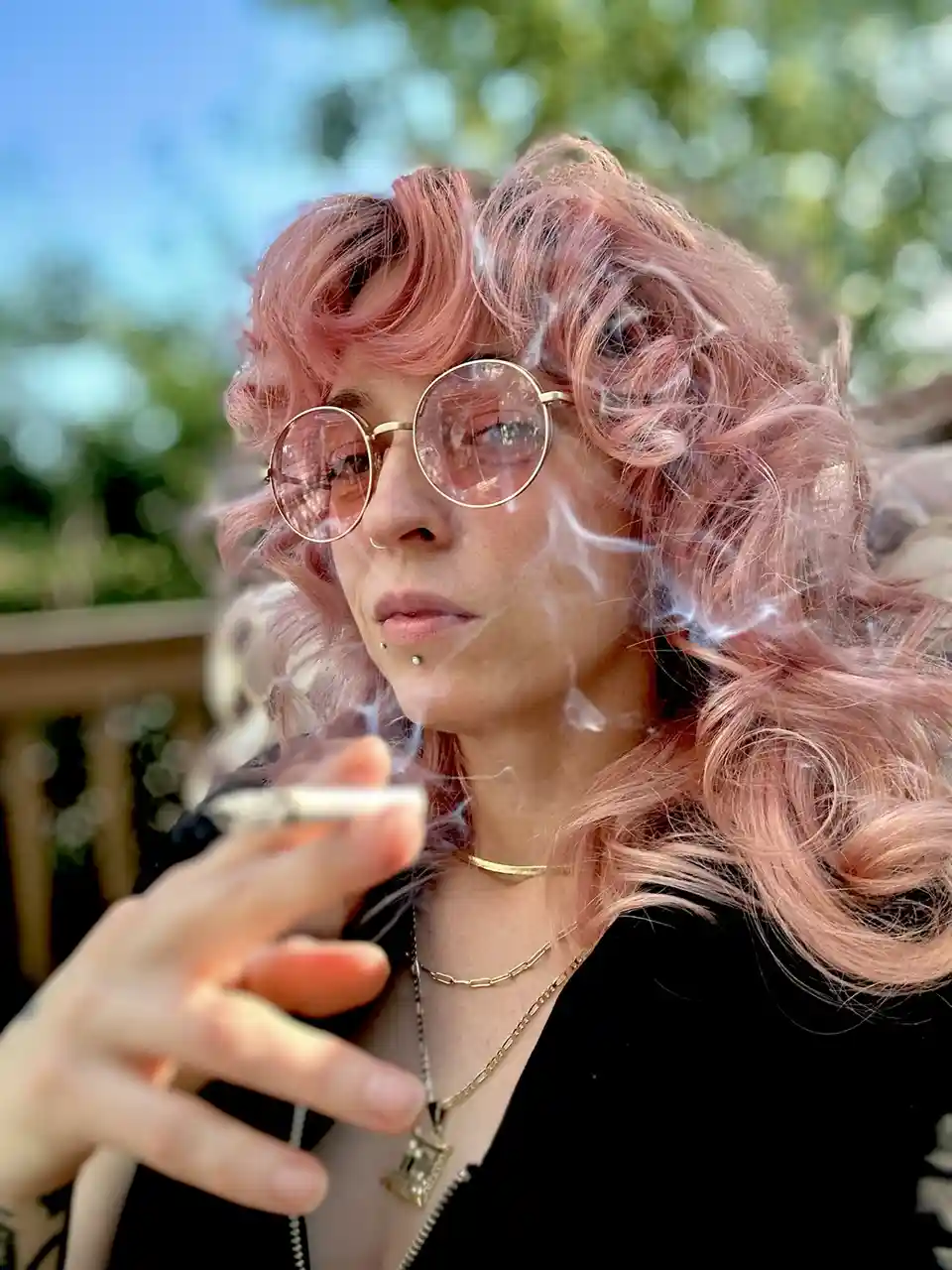 A person with curly pink hair and round gold-rimmed glasses exhales smoke while holding a cigarette outdoors. They wear…