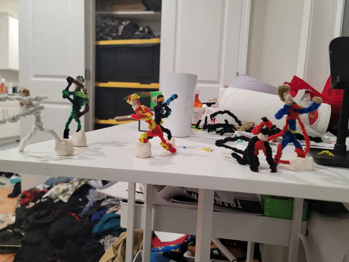 A cluttered white table holds several handmade pipe cleaner figures resembling superheroes, each standing on a small clay…