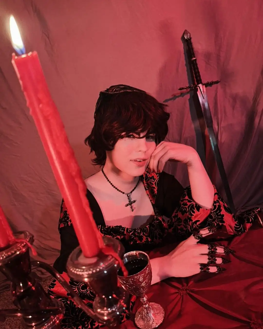 A person with short dark hair, wearing a black and red patterned dress with a cross necklace, sits in a dimly lit red room…