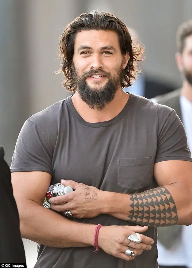 a man with a beard and tattoos on his arms, wearing a grey t-shirt and holding a bottle, smiles while looking off to the…
