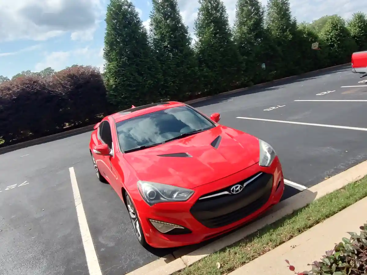 A bright red Hyundai Genesis Coupe is parked at an angle in a paved parking lot, partially over the curb and sidewalk. The…
