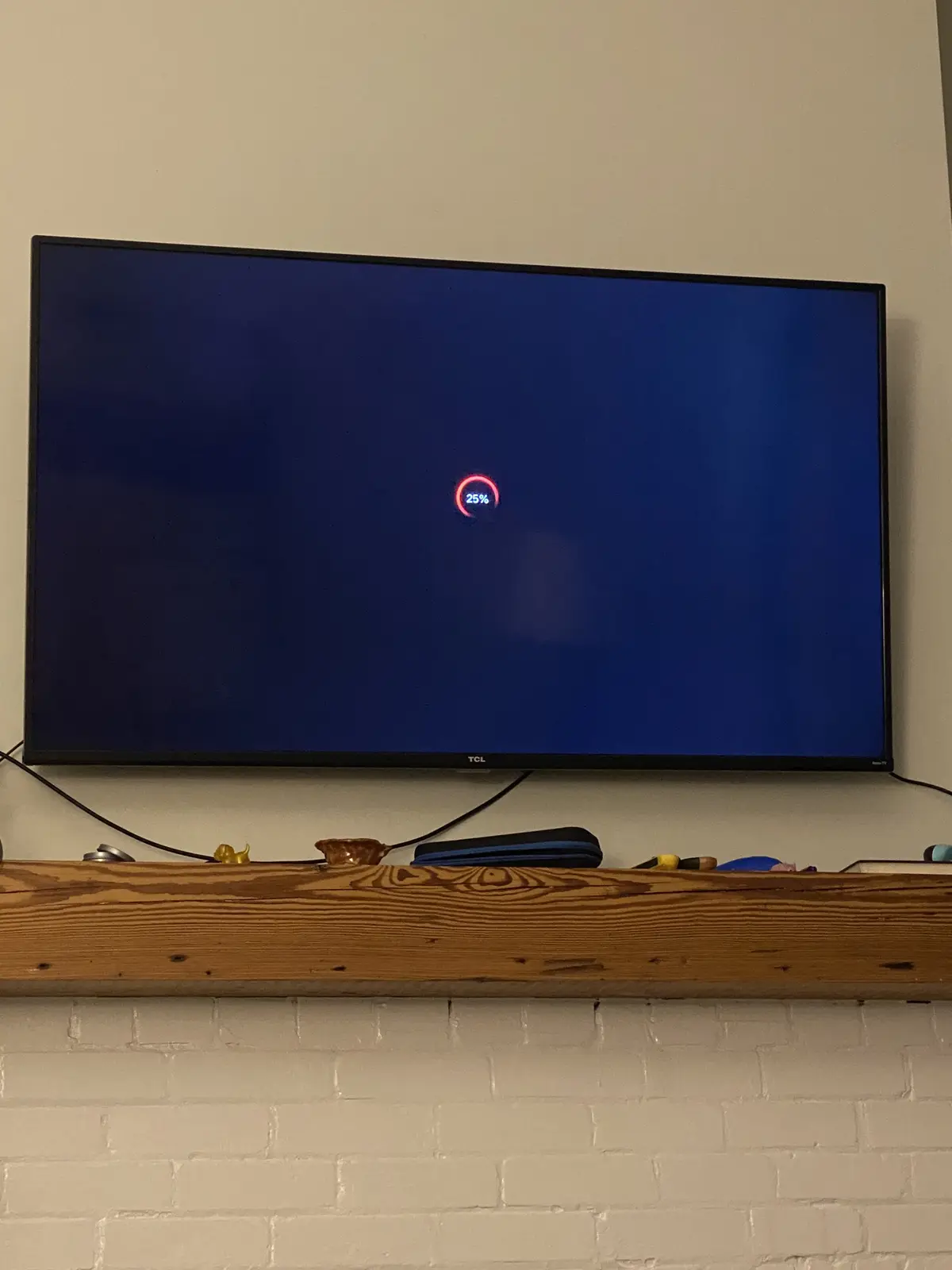 A TCL TV mounted on a wall above a wooden beam shows a blue screen with a red loading circle and the text '25%' in the…