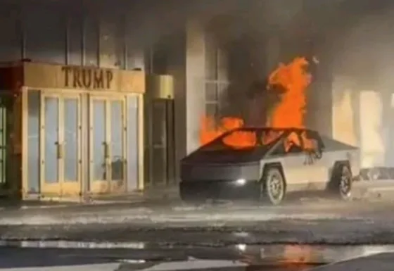 A Tesla Cybertruck is on fire in front of a building with a sign that reads 'TRUMP'. The vehicle is engulfed in flames and…