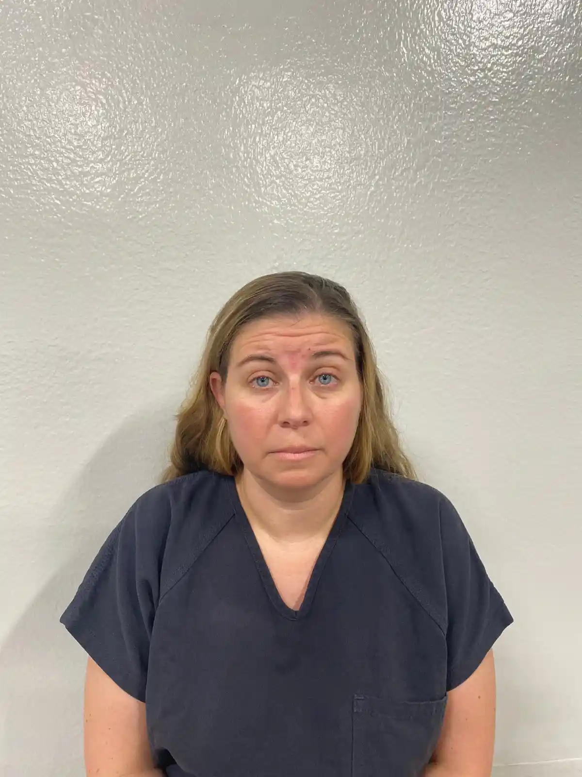 A woman with blonde hair and blue eyes, wearing dark blue scrubs, stares directly at the camera with a tired, unimpressed…