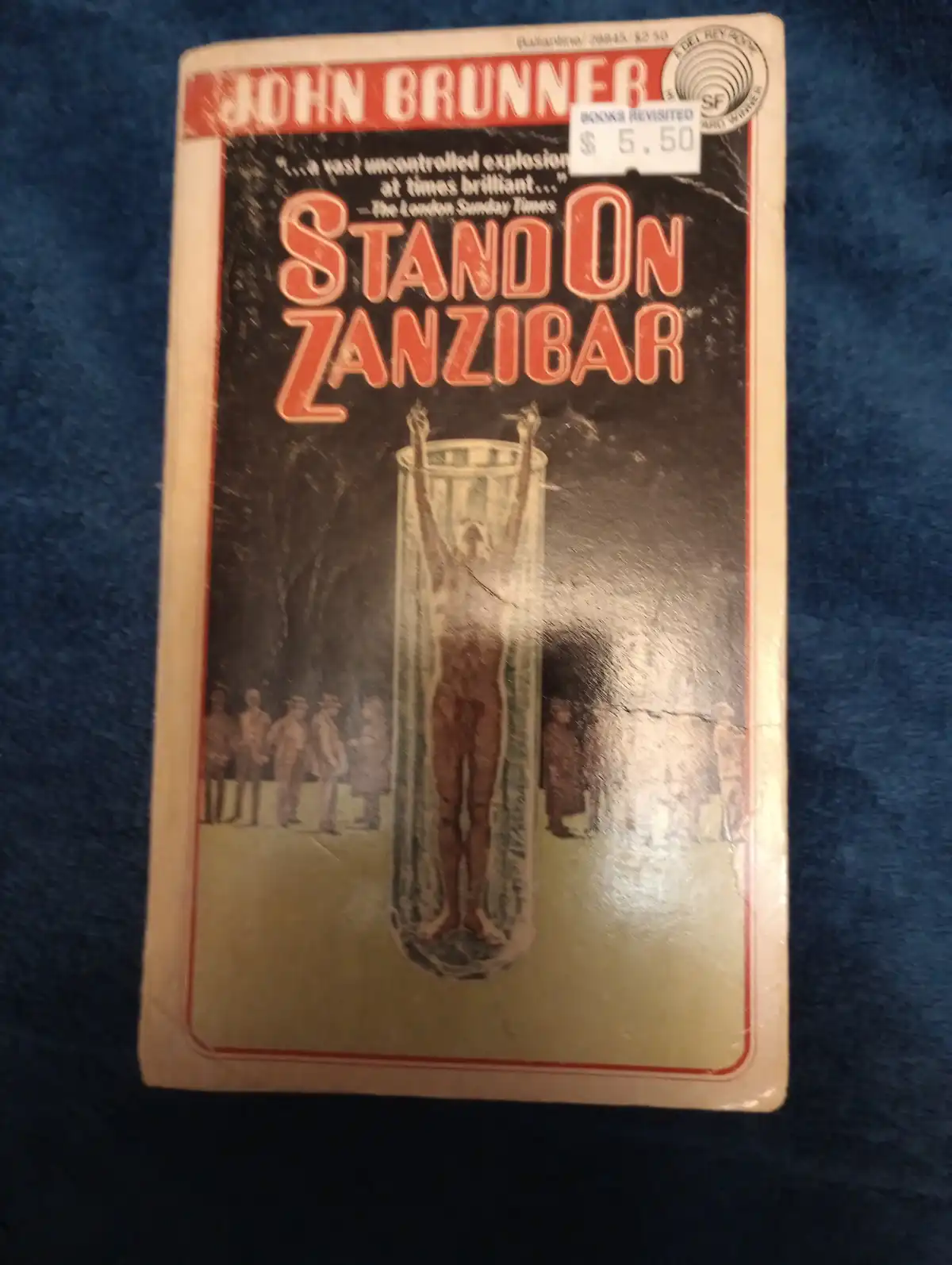 A vintage paperback book cover for John Brunner's 'Stand on Zanzibar' shows a person standing inside a giant glass test tube…