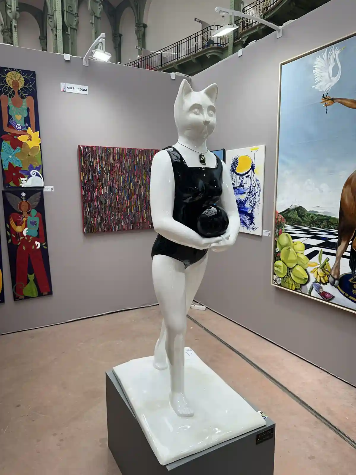 A white ceramic statue of a cat with a human body, wearing a black leotard and holding a black ball, stands on a pedestal in…
