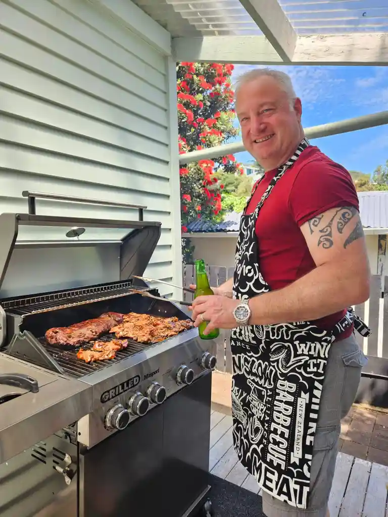 A smiling man in a red shirt and a black-and-white barbecue-themed apron stands beside a large stainless steel grill…
