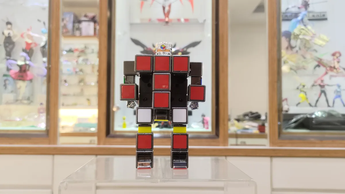 A robot figure constructed from Rubik's cube pieces stands on a clear display stand, with a background of glass display…