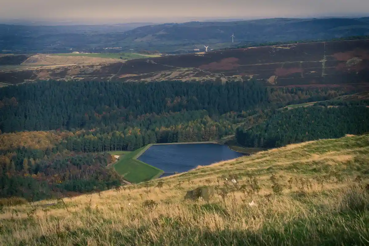 A scenic landscape photo shows a large reservoir nestled among rolling hills, forests, and distant wind turbines under a…