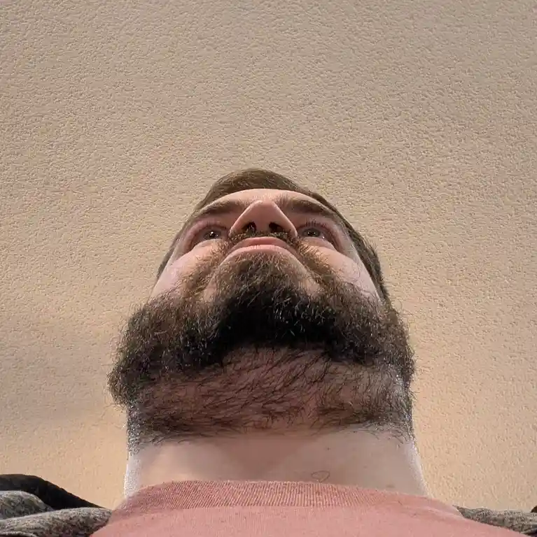 A low-angle shot shows a man with a full beard and mustache looking upward, his eyes directed toward the ceiling. He wears a…