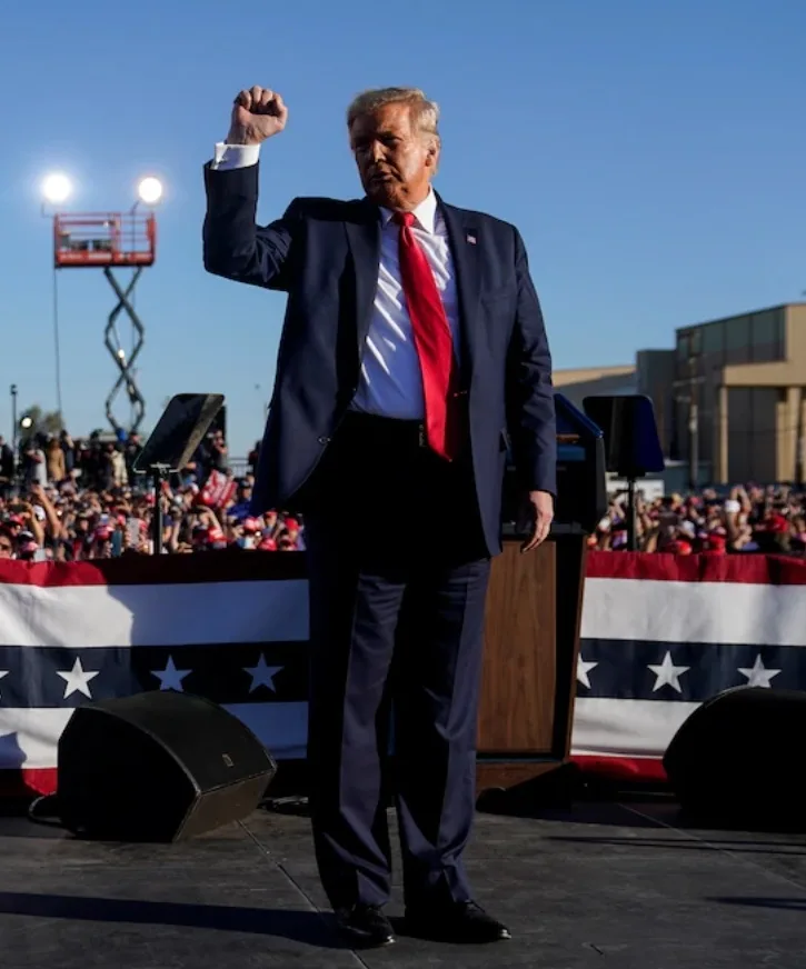 a man in a dark suit and red tie stands on a stage with his right fist raised, facing slightly away from the camera. Behind…