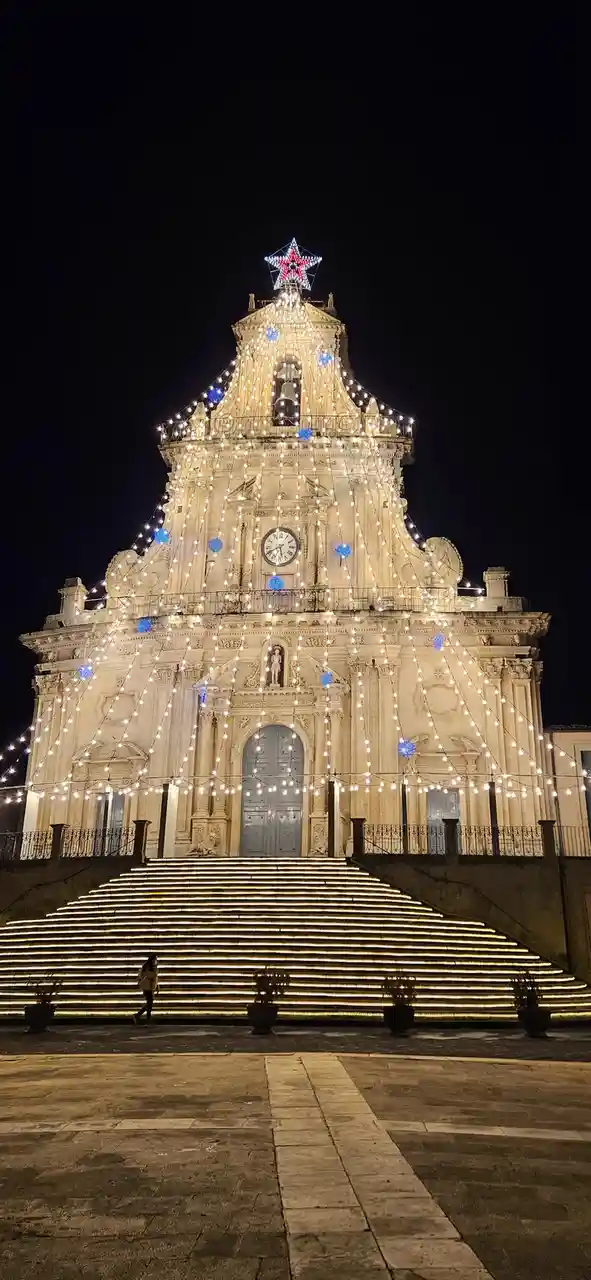 a grand, ornate church facade at night is festively decorated with strings of warm white lights and blue spherical…