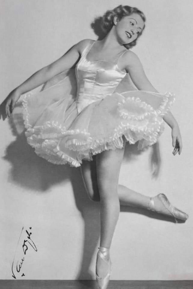 A vintage black-and-white photograph shows a smiling woman in a classic ballet tutu and pointe shoes striking a graceful…
