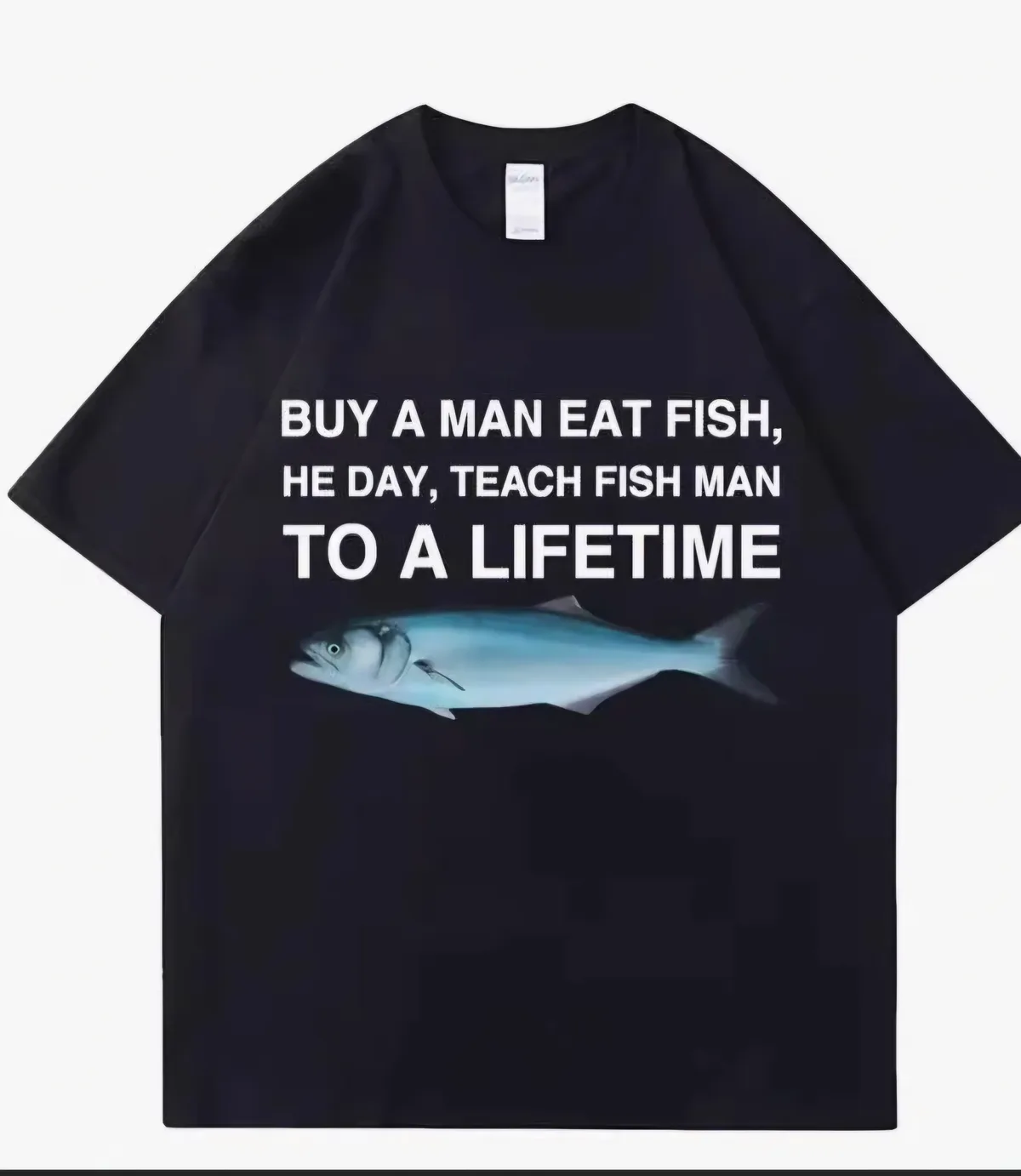 A black t-shirt displays a humorous, grammatically incorrect phrase in bold white text: 'BUY A MAN EAT FISH, HE DAY, TEACH…