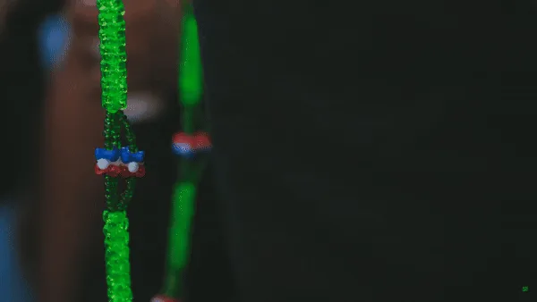 The image shows a close-up of a green beaded lanyard with a small, colorful charm shaped like a face with blue eyes, red…