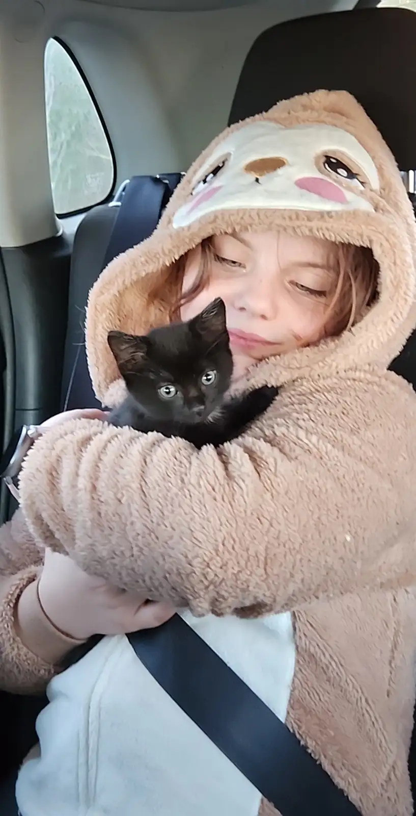 A young person wearing a fluffy, tan animal-themed hooded onesie sits in a car seat, holding a small black kitten close to…