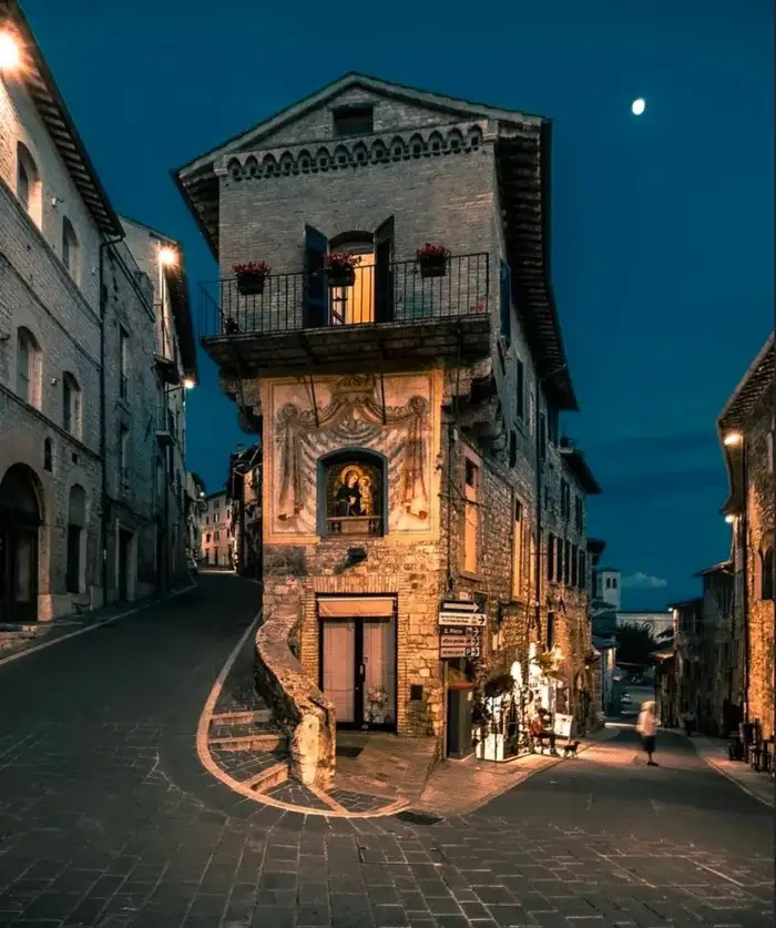 A picturesque European street at dusk, featuring a stone building with a balcony and a religious fresco, illuminated by warm…